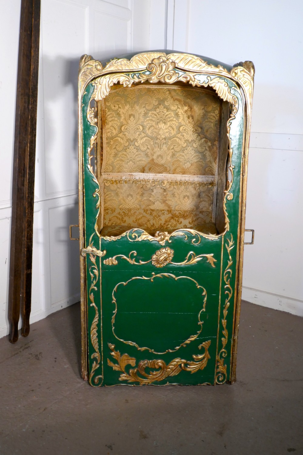 A 19th-century green painted and giltwood Italian sedan chair featuring hand-carved scrollwork, window openings, gold fabric lining, and wooden carrying poles. View V