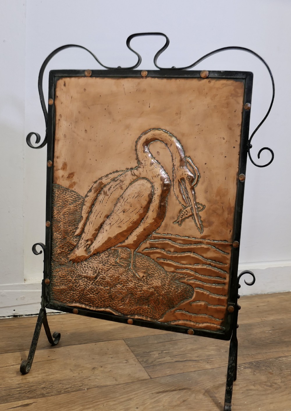 An Arts and Crafts wrought iron fire screen with a hammered copper panel depicting a repousse stork catching a fish near water.