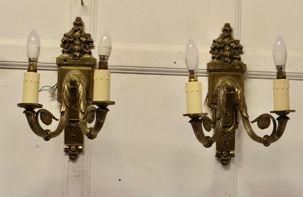 A pair of Neoclassical cast brass twin-arm wall lights with decorative backplates, acanthus leaf details, and candle sleeves.