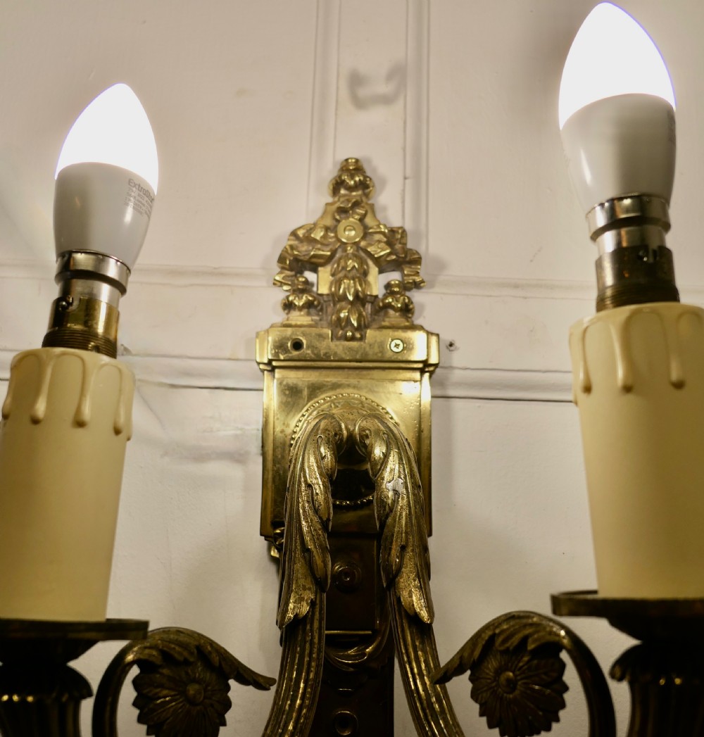 A pair of Neoclassical cast brass twin-arm wall lights with decorative backplates, acanthus leaf details, and candle sleeves. View IX