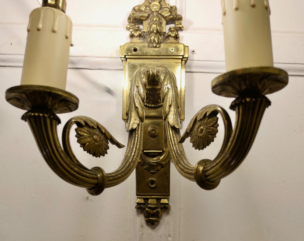 A pair of Neoclassical cast brass twin-arm wall lights with decorative backplates, acanthus leaf details, and candle sleeves. View X