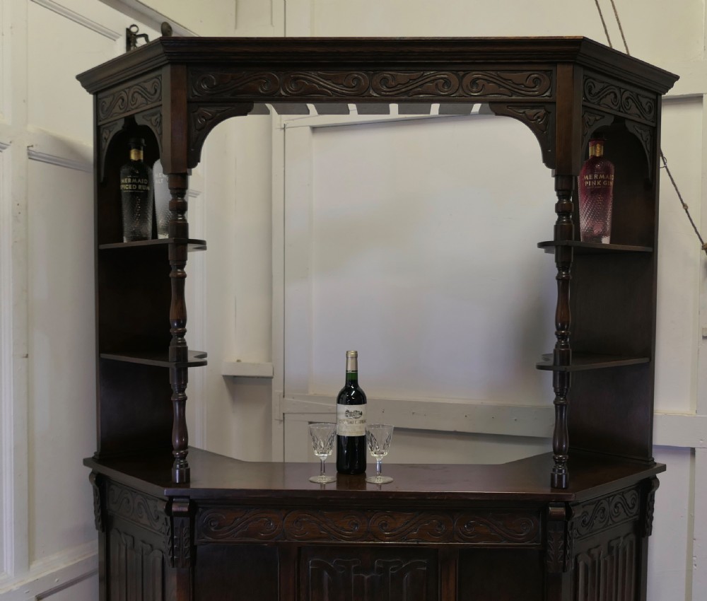A dark brown carved oak canopy bar featuring turned spindle columns, open side shelves, an overhead frieze, and vertical panelled detailing on the base. View V
