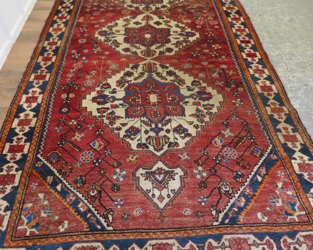 A large red wool carpet featuring traditional central medallion designs, geometric floral motifs, and a navy blue decorative border. View II