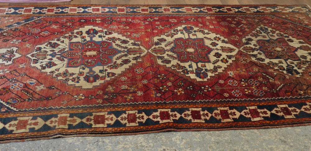 A large red wool carpet featuring traditional central medallion designs, geometric floral motifs, and a navy blue decorative border. View III