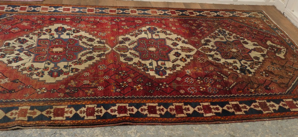 A large red wool carpet featuring traditional central medallion designs, geometric floral motifs, and a navy blue decorative border. View V