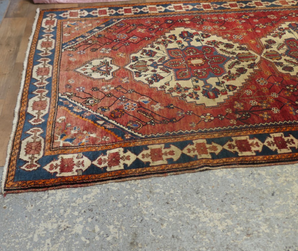 A large red wool carpet featuring traditional central medallion designs, geometric floral motifs, and a navy blue decorative border. View VI