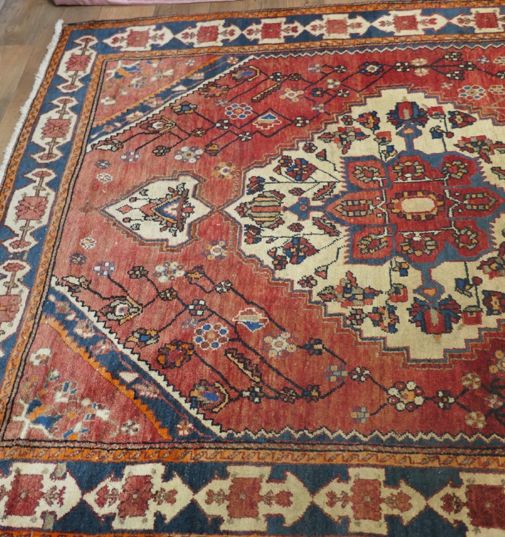 A large red wool carpet featuring traditional central medallion designs, geometric floral motifs, and a navy blue decorative border. View IX