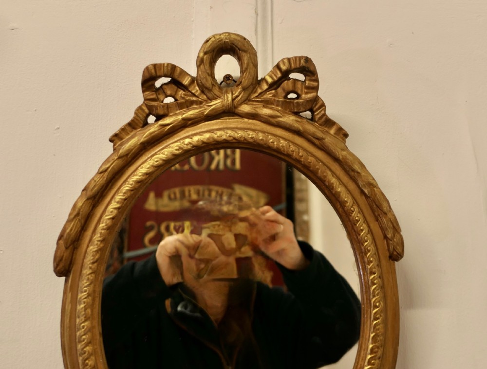 A French Louis XVI style oval giltwood wall mirror featuring a carved ribbon bow cresting and gold leaf finish. View IV