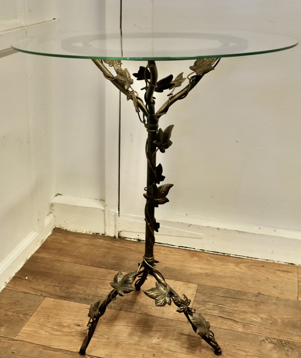 A small circular glass-topped wine table resting on a dark bronze metal tripod base sculpted into trailing ivy vine leaves. View VI