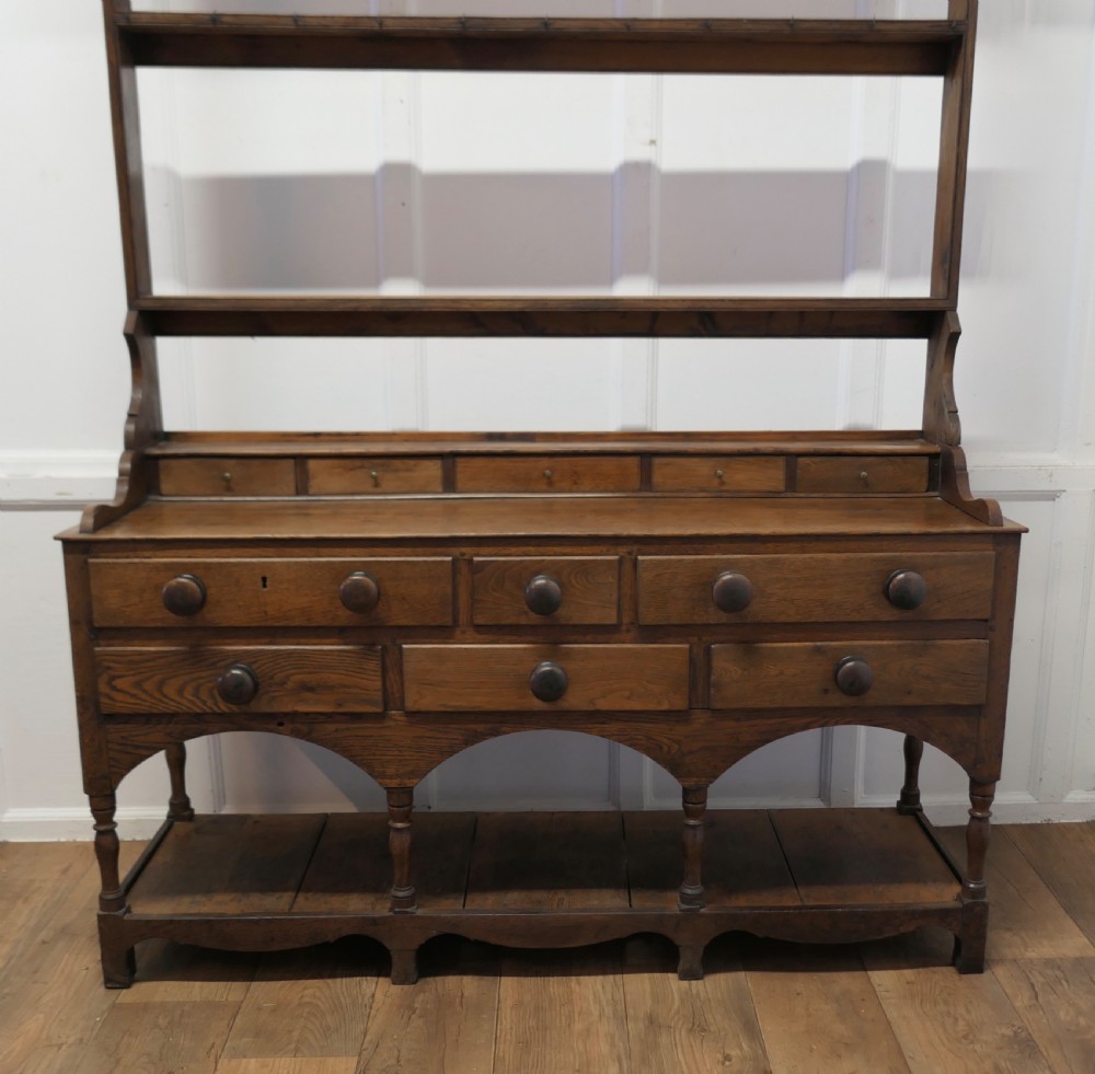 A Georgian country oak dresser featuring an open tiered plate rack set above multiple drawer compartments, arched aprons, and an open pot shelf at the base.