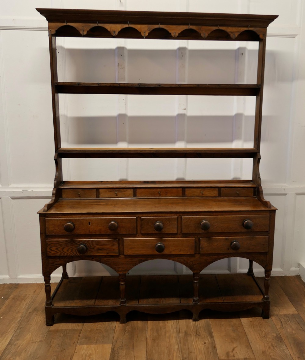 A Georgian country oak dresser featuring an open tiered plate rack set above multiple drawer compartments, arched aprons, and an open pot shelf at the base. View II