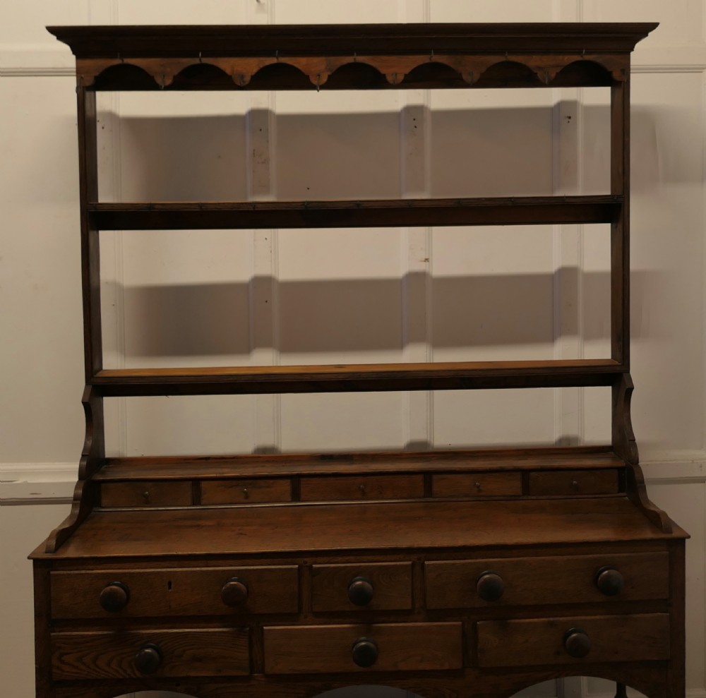 A Georgian country oak dresser featuring an open tiered plate rack set above multiple drawer compartments, arched aprons, and an open pot shelf at the base. View III