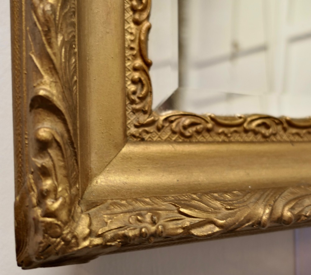 A rectangular antique giltwood wall mirror featuring ornate relief foliage moulding and a bevelled glass plate. View VI
