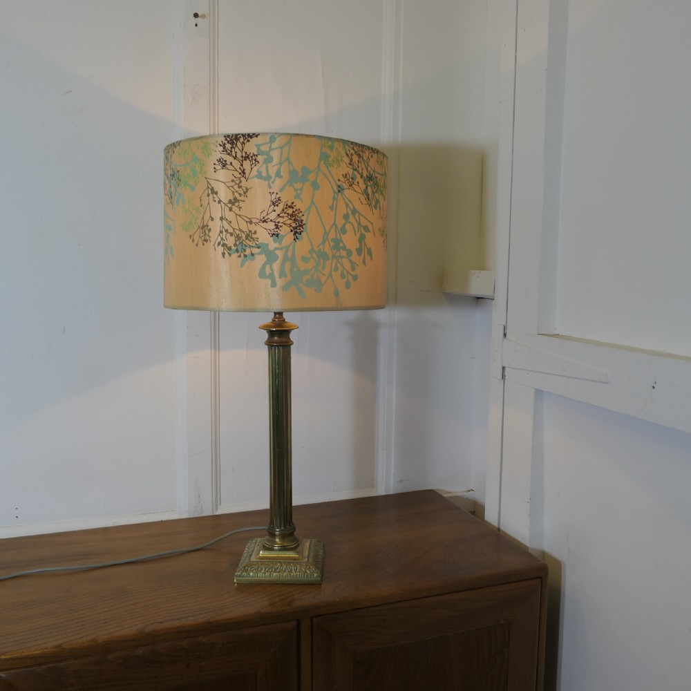 A tall brass table lamp featuring a fluted column stem on a stepped square base, topped with a cylindrical cream shade patterned with botanical motifs.
