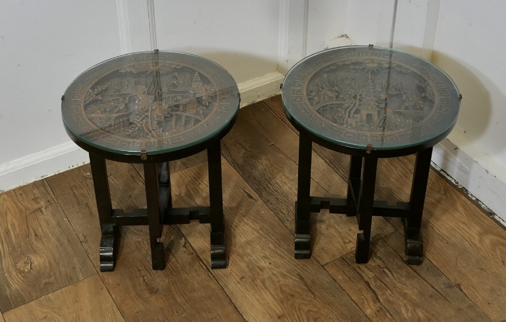 A pair of circular dark wood side tables featuring relief-carved landscape panels beneath fitted glass tops.