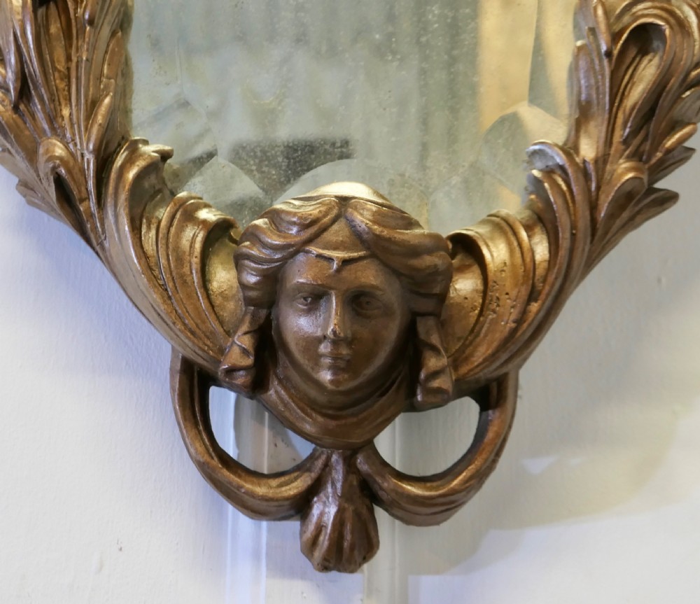 An oval antique giltwood wall mirror framed by intricately carved cherubs, acanthus scrollwork, and a sculpted mask base. View VII