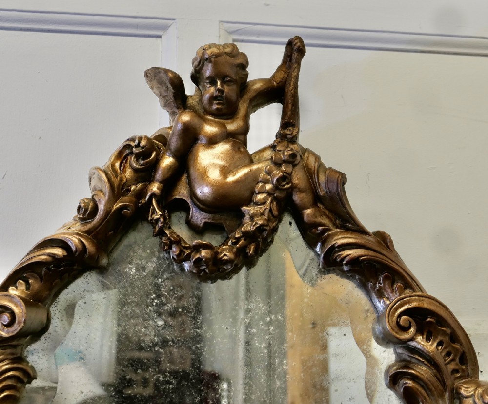An oval antique giltwood wall mirror framed by intricately carved cherubs, acanthus scrollwork, and a sculpted mask base. View VIII