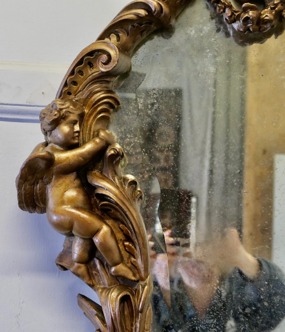 An oval antique giltwood wall mirror framed by intricately carved cherubs, acanthus scrollwork, and a sculpted mask base. View IX