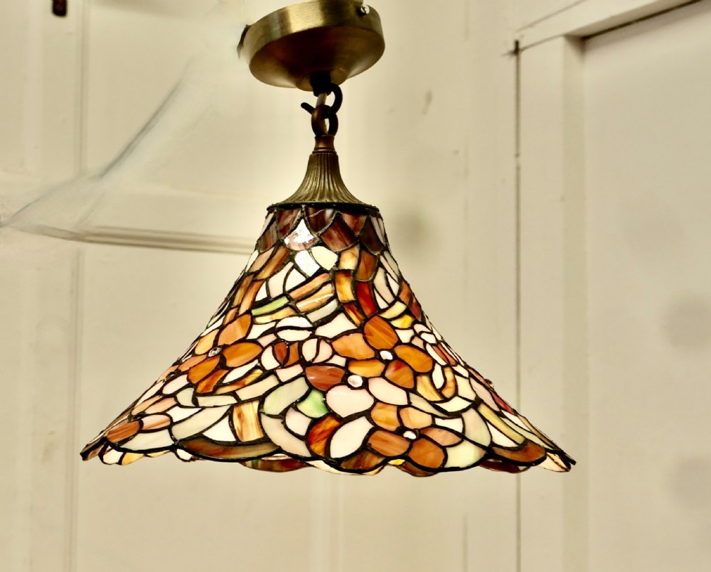 A conical Arts and Crafts stained glass lampshade featuring an amber and cream floral pattern with a scalloped lower edge and brass ceiling fittings.