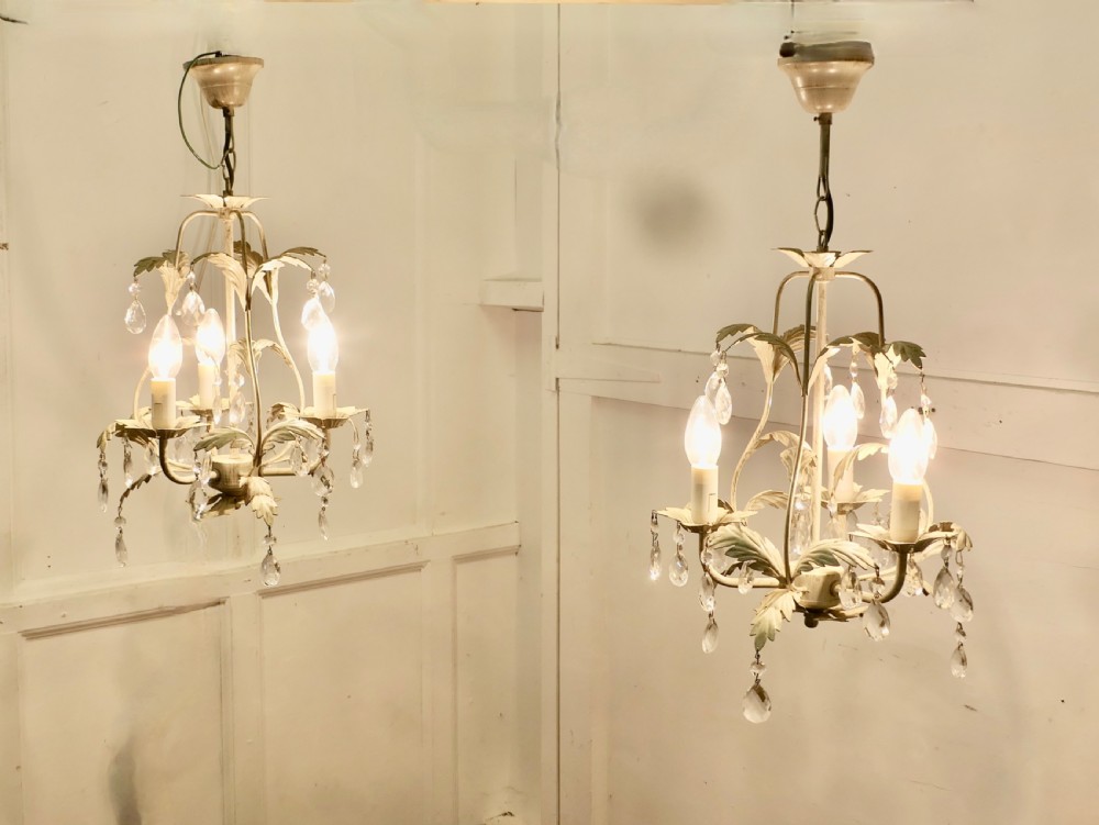 A pair of white painted metal open basket chandeliers featuring foliate leaf motifs, candle bulb holders, and suspended glass droplets.