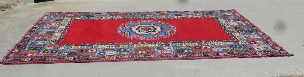 A red hand-knotted wool rug featuring a large central blue and cream star medallion surrounded by an intricate multi-coloured geometric border. View II