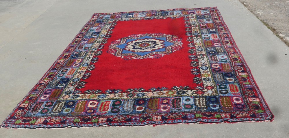 A red hand-knotted wool rug featuring a large central blue and cream star medallion surrounded by an intricate multi-coloured geometric border. View III