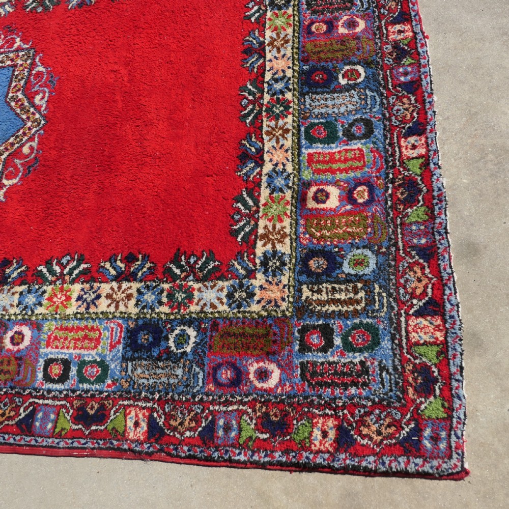 A red hand-knotted wool rug featuring a large central blue and cream star medallion surrounded by an intricate multi-coloured geometric border. View V