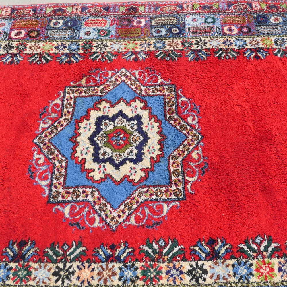 A red hand-knotted wool rug featuring a large central blue and cream star medallion surrounded by an intricate multi-coloured geometric border. View VI
