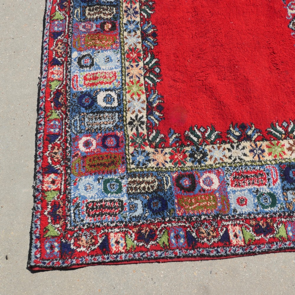 A red hand-knotted wool rug featuring a large central blue and cream star medallion surrounded by an intricate multi-coloured geometric border. View VII