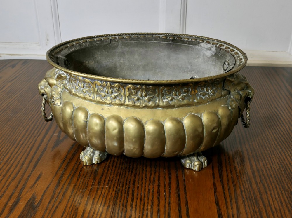 An oval antique brass jardiniere featuring gadrooned lower detail, lion mask handles with drop rings, paw feet, and a removable internal metal liner.