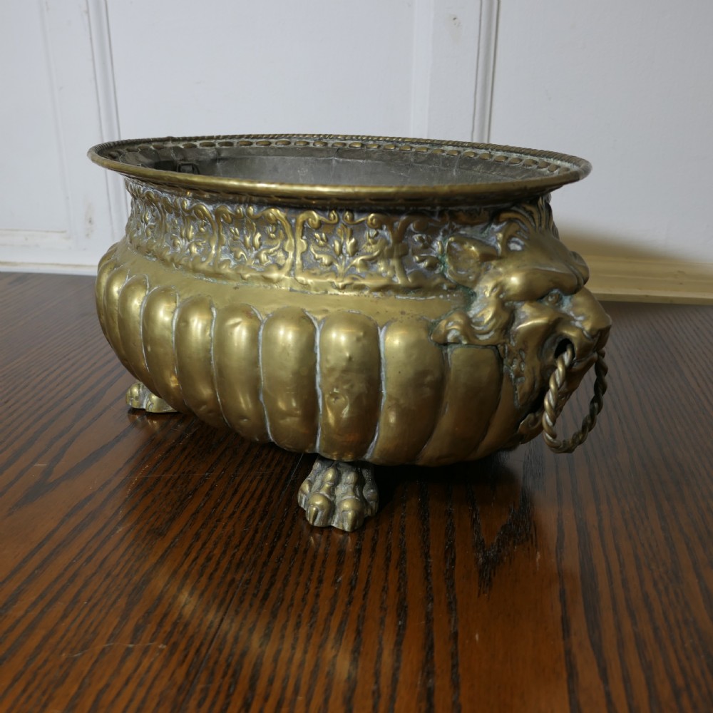 An oval antique brass jardiniere featuring gadrooned lower detail, lion mask handles with drop rings, paw feet, and a removable internal metal liner. View V