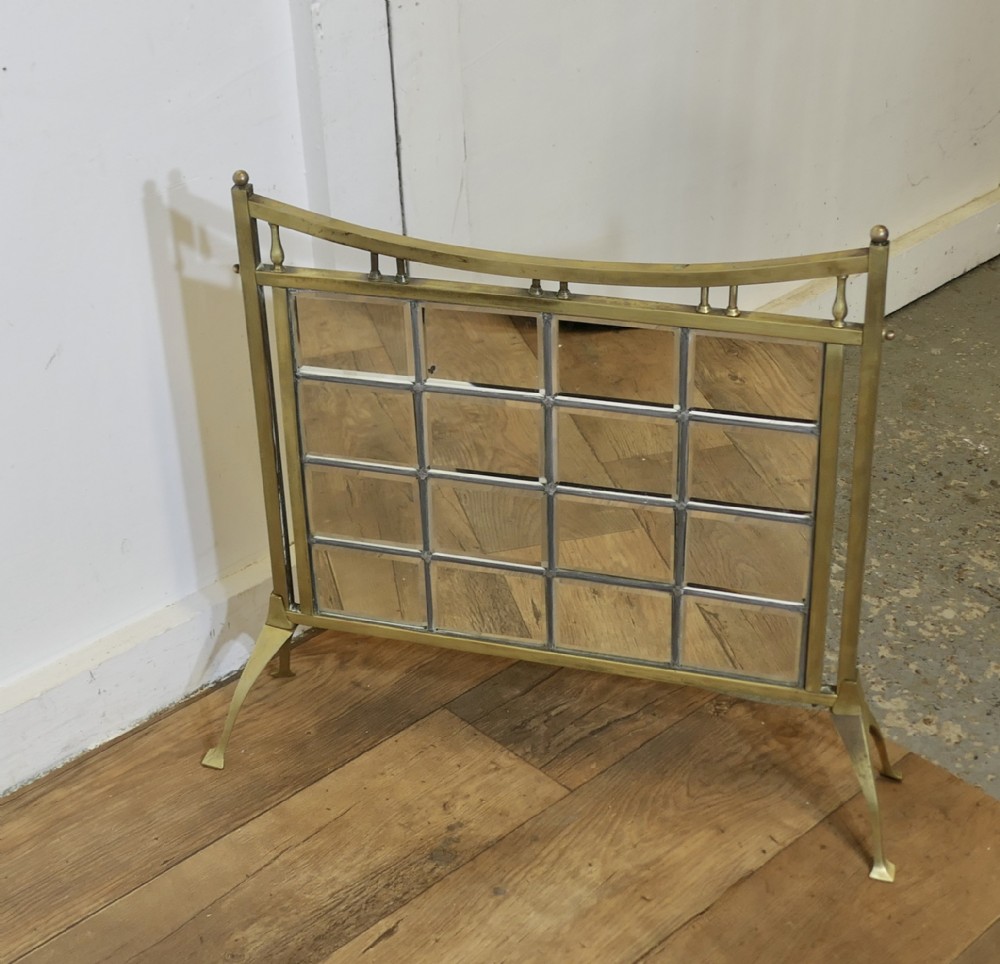 An Arts and Crafts style brass fire screen featuring a curved top rail with vertical spindles above a panel of fifteen rectangular mirrored panes resting on splayed feet.