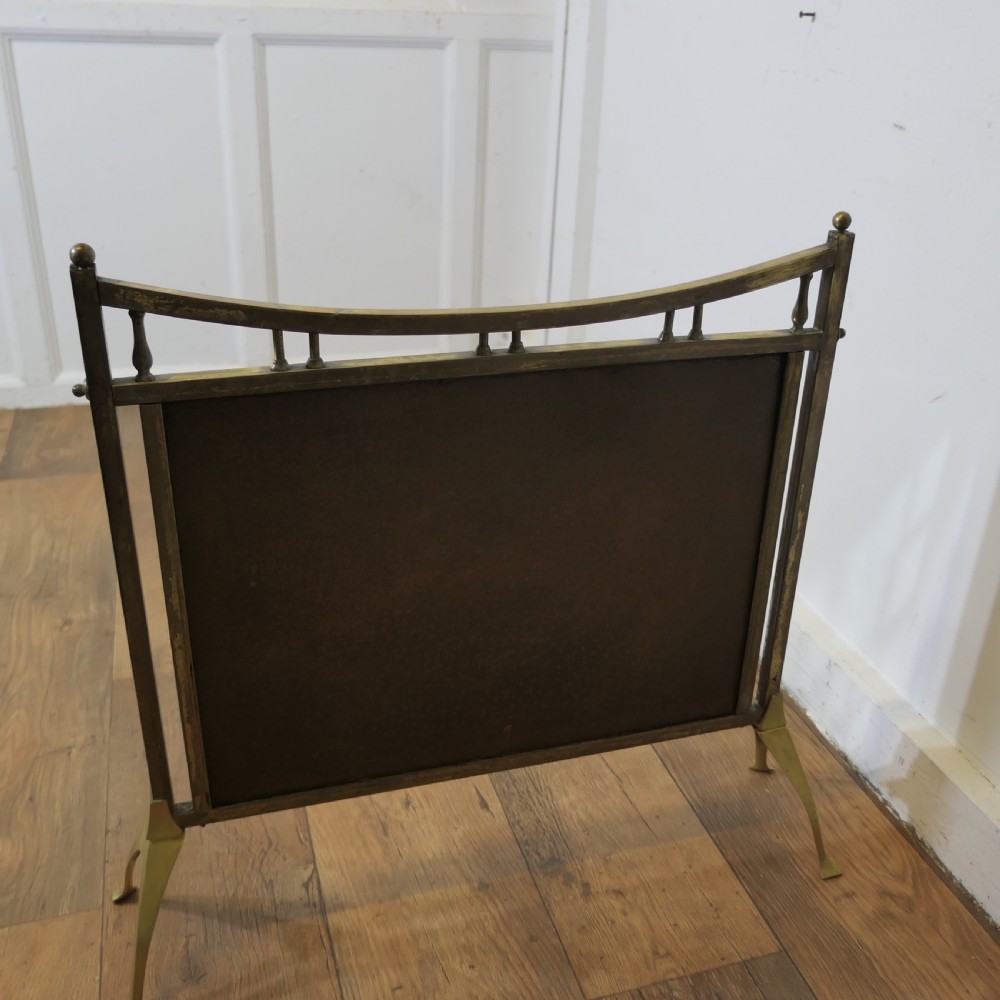 An Arts and Crafts style brass fire screen featuring a curved top rail with vertical spindles above a panel of fifteen rectangular mirrored panes resting on splayed feet. View VI