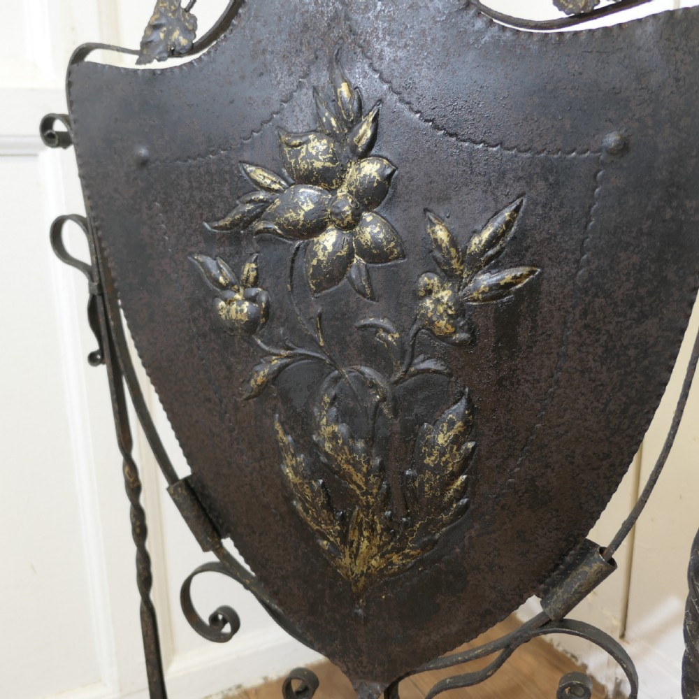 A black wrought iron fire screen with a shield-shaped central panel featuring gilded floral repousse detailing and scrollwork legs. View V