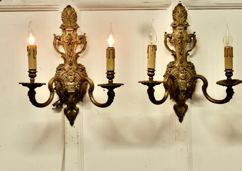 A pair of ornate cast brass twin-arm wall sconces featuring Rococo scrollwork and faux candle drip sleeves.
