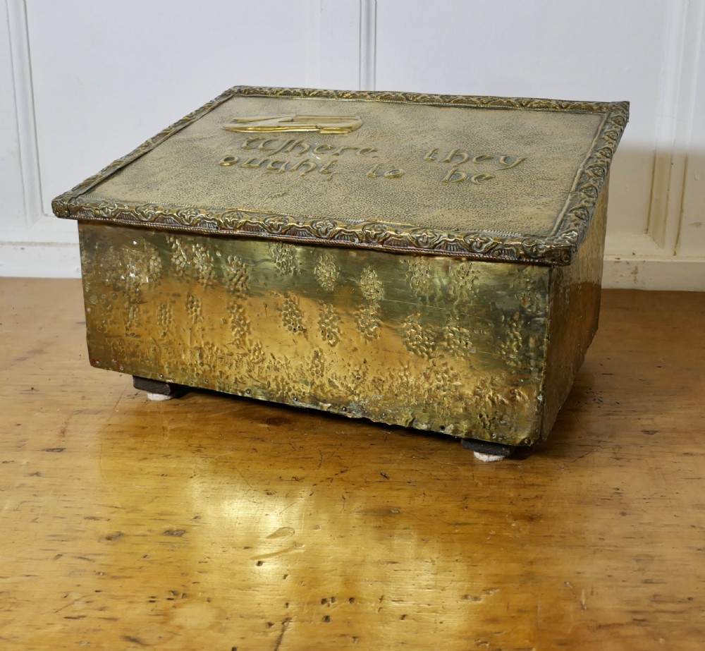 A hand-beaten brass slipper box featuring a sloped lid embossed with footwear imagery and decorative lettering reading Where they ought to be.