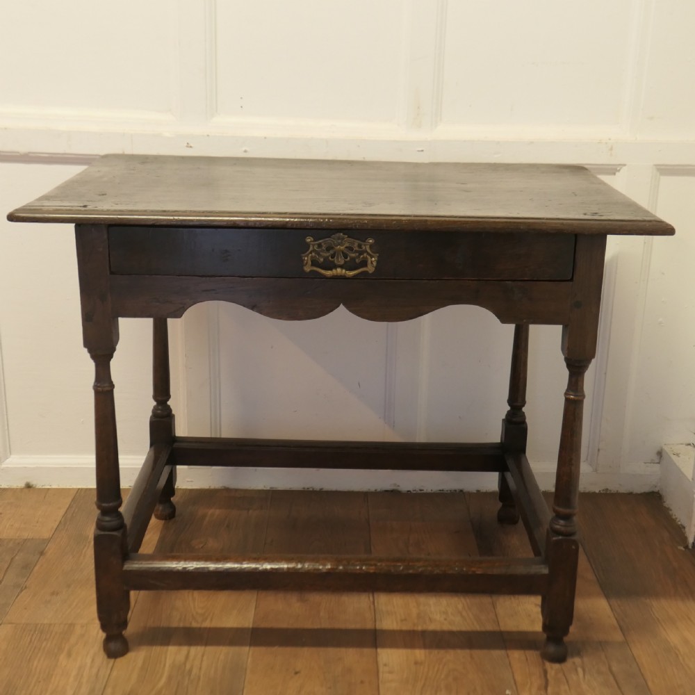 An eighteenth-century dark oak lowboy table featuring a single drawer with a brass handle, a shaped lower apron, turned legs, and a perimeter stretcher.