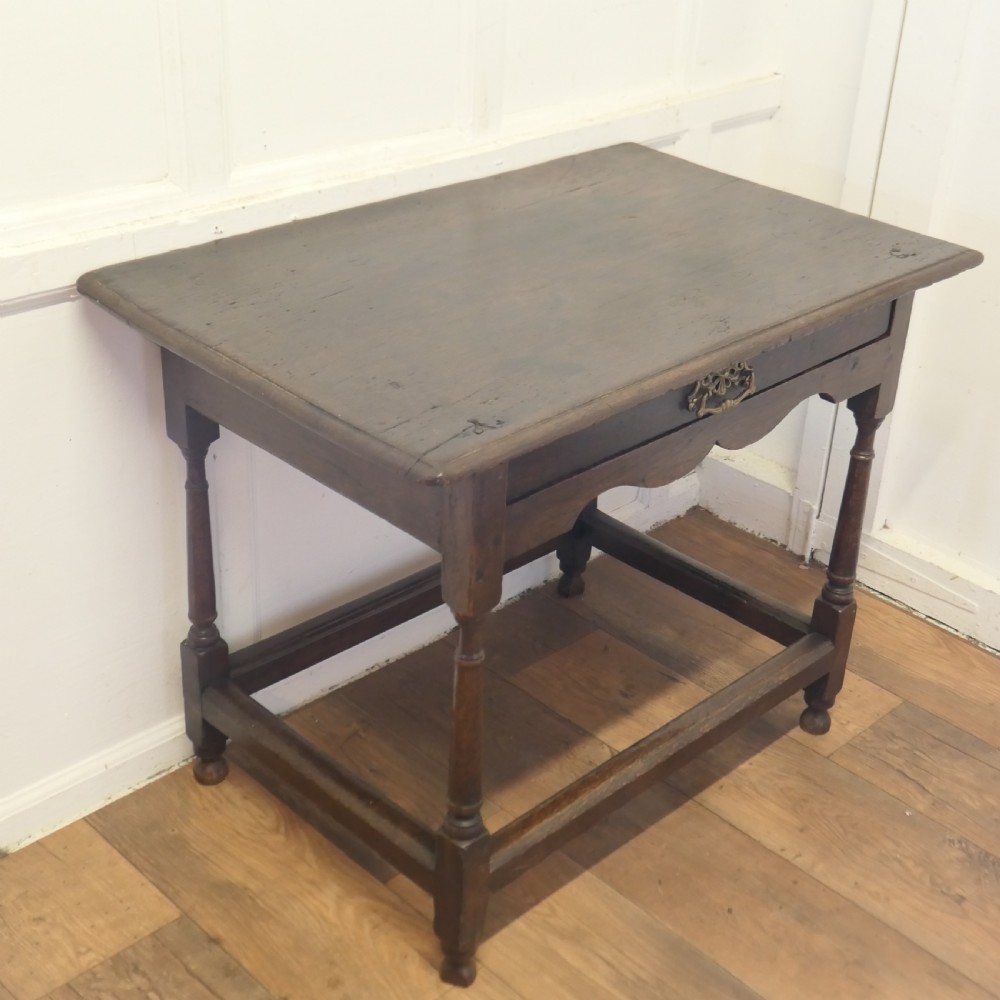 An eighteenth-century dark oak lowboy table featuring a single drawer with a brass handle, a shaped lower apron, turned legs, and a perimeter stretcher. View III