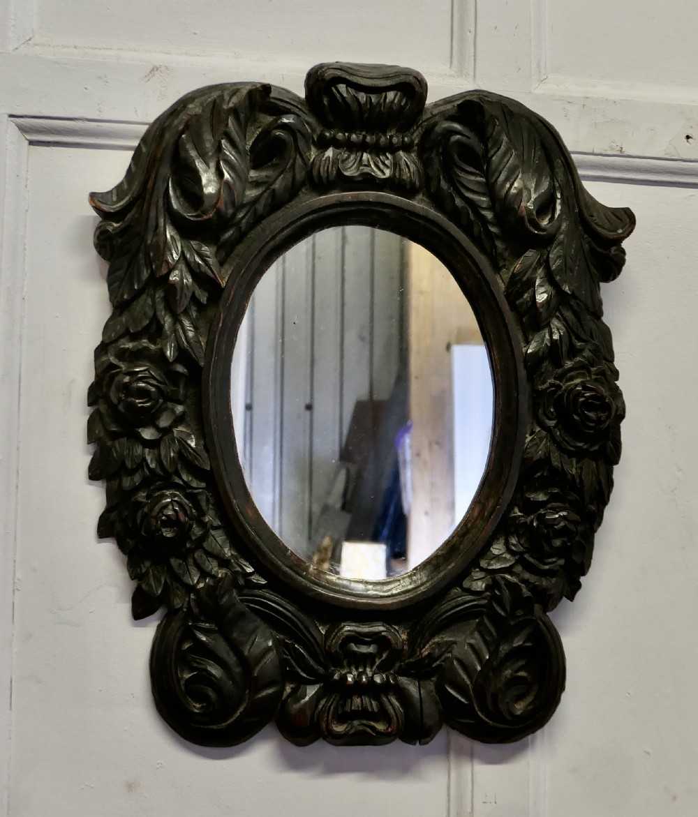 An oval glass mirror enclosed within a deeply carved dark oak frame featuring floral and leaf relief details.
