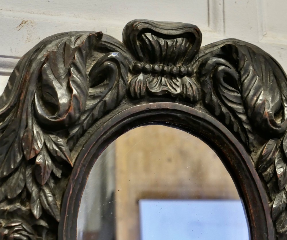 An oval glass mirror enclosed within a deeply carved dark oak frame featuring floral and leaf relief details. View III