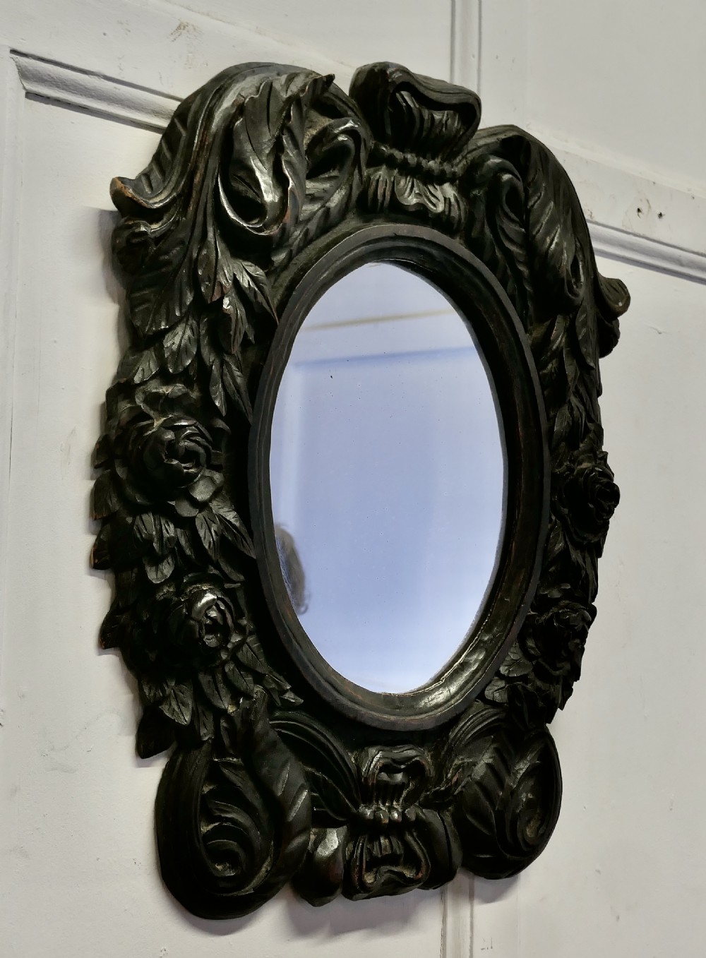 An oval glass mirror enclosed within a deeply carved dark oak frame featuring floral and leaf relief details. View V