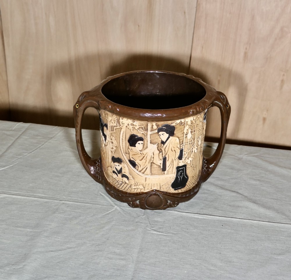 A cylindrical Bretby earthenware jardiniere featuring dark brown glazed twin handles and a cream central band decorated with relief-carved Japanese figures.