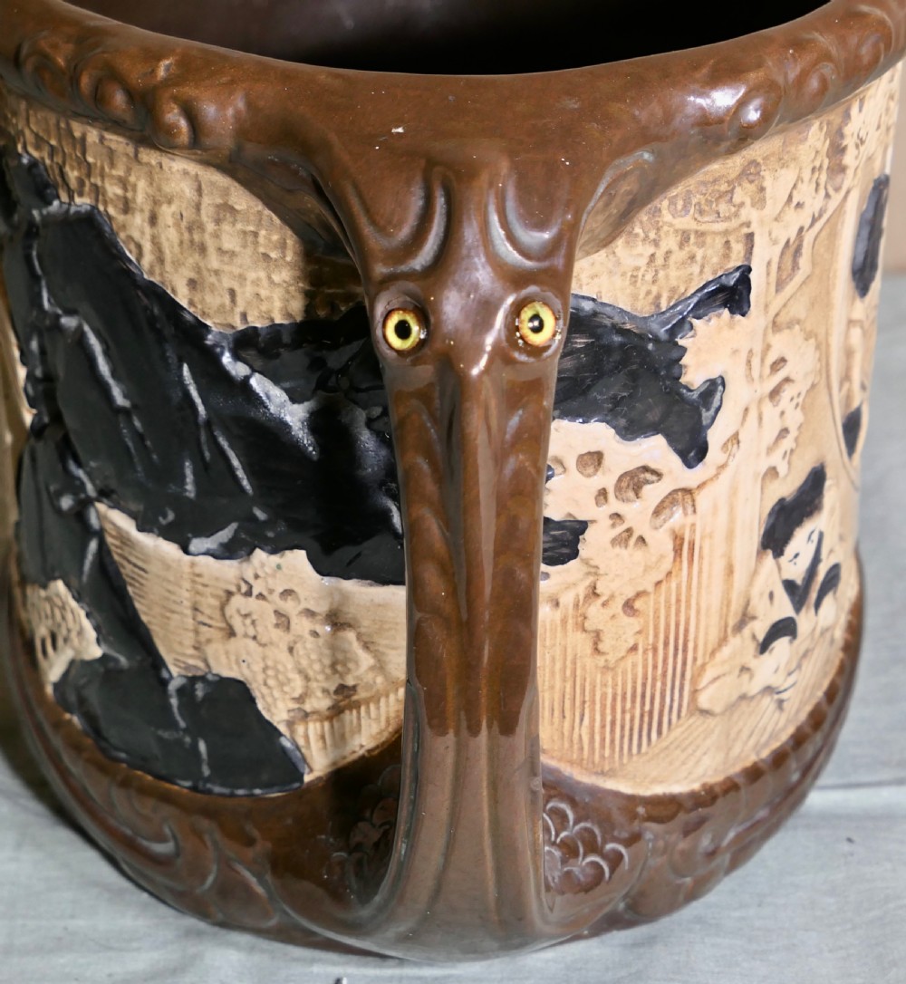 A cylindrical Bretby earthenware jardiniere featuring dark brown glazed twin handles and a cream central band decorated with relief-carved Japanese figures. View IX