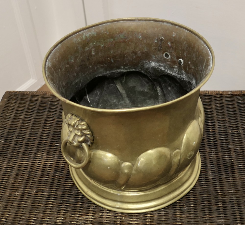A large round brass jardiniere with gadrooned lower relief, flared upper rim, stepped base, and cast lion mask handles holding drop rings. View II