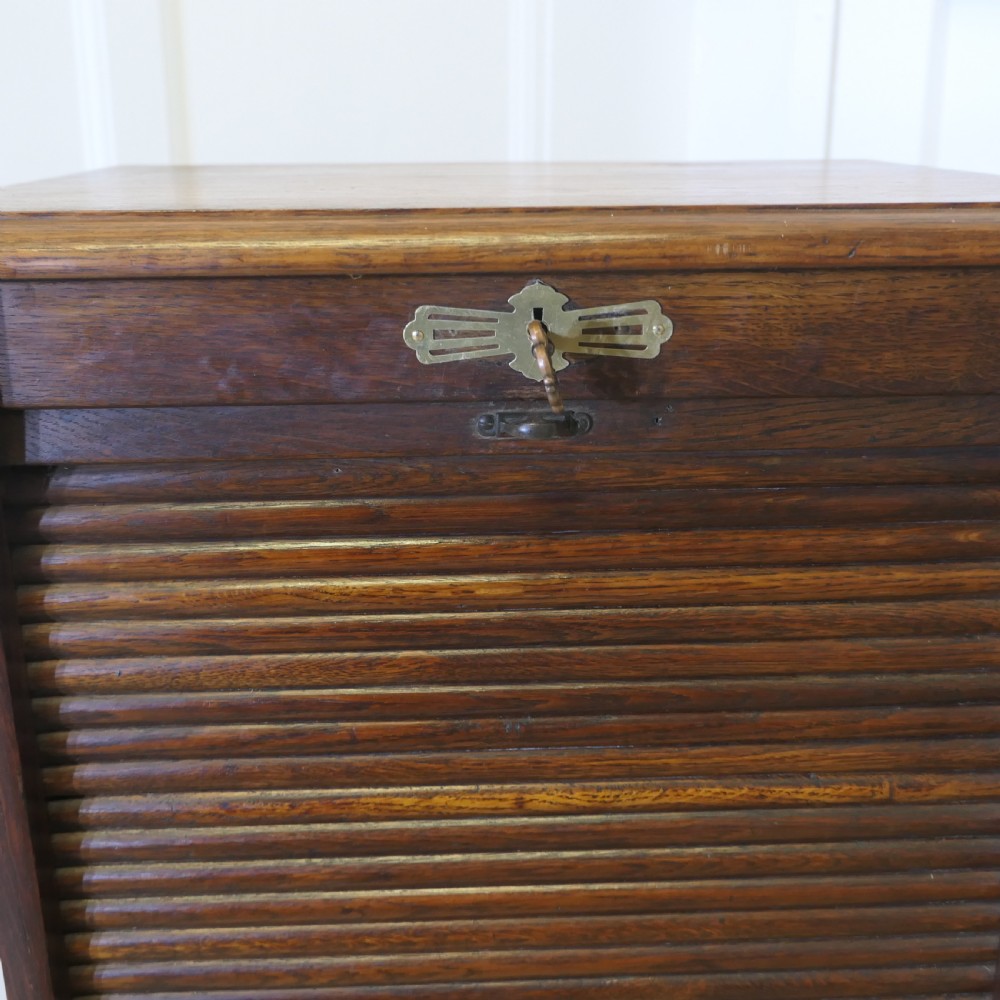 A tall medium brown oak filing cabinet featuring a flexible slatted tambour front, top keyhole escutcheon plate, and a solid plinth base. View IV