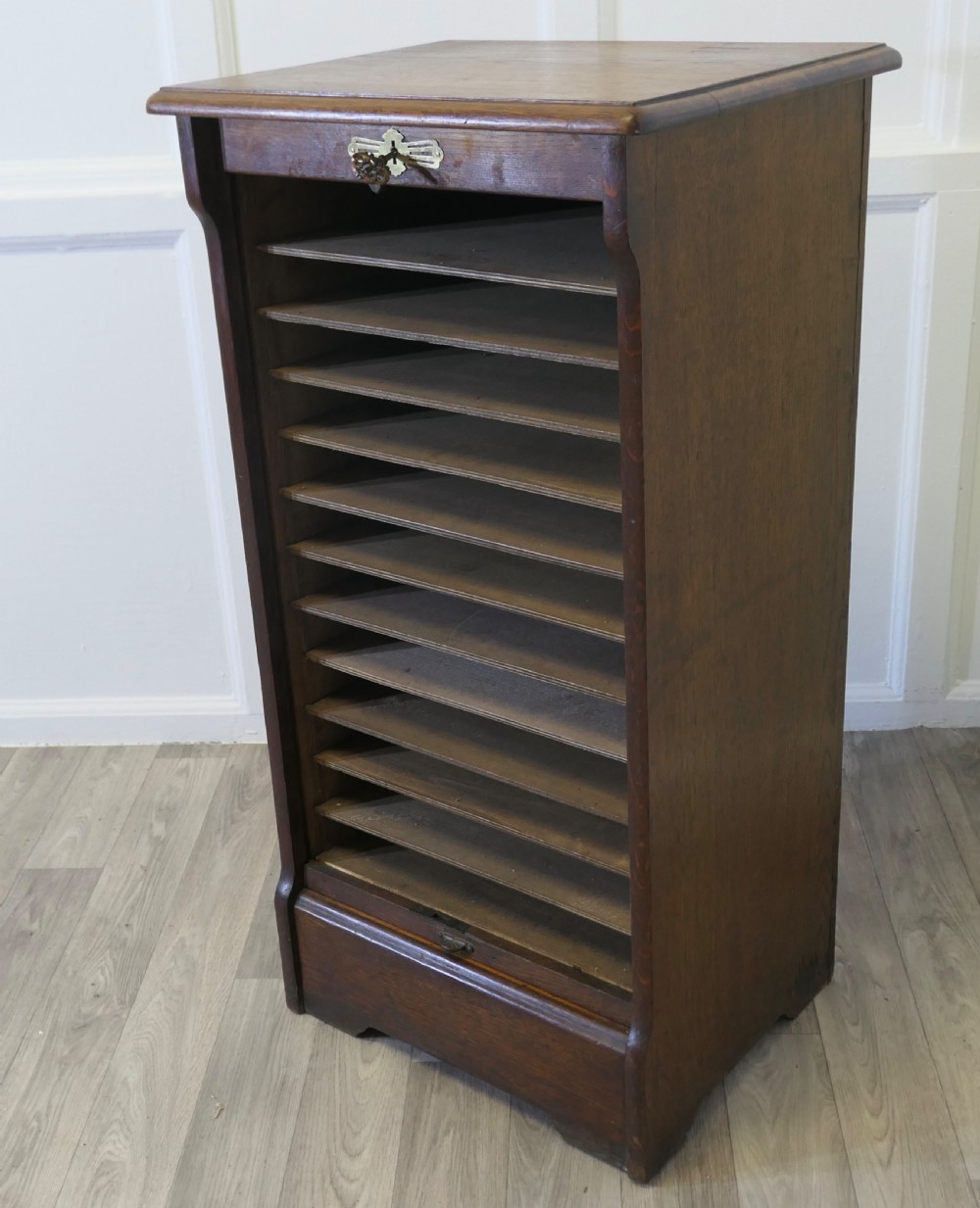 A tall medium brown oak filing cabinet featuring a flexible slatted tambour front, top keyhole escutcheon plate, and a solid plinth base. View V