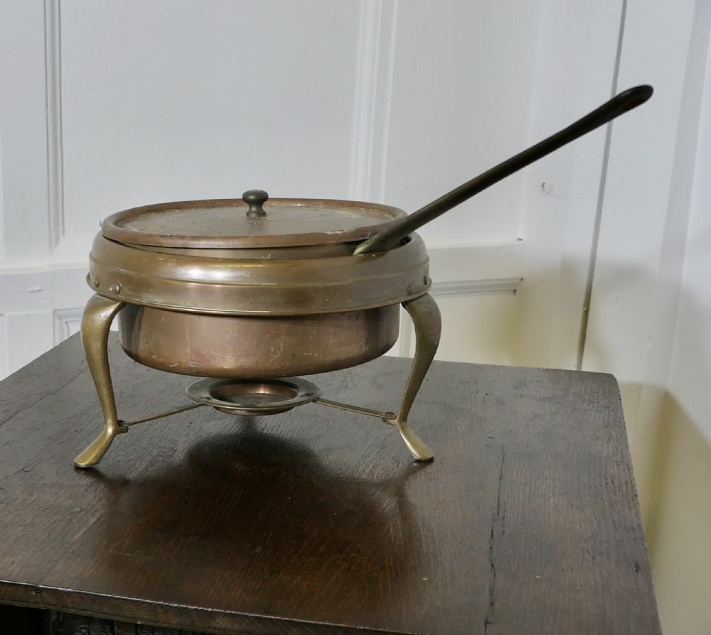 A vintage circular copper bain marie featuring a lid, long handle, and a three-legged brass tripod stand with an integrated burner tray.