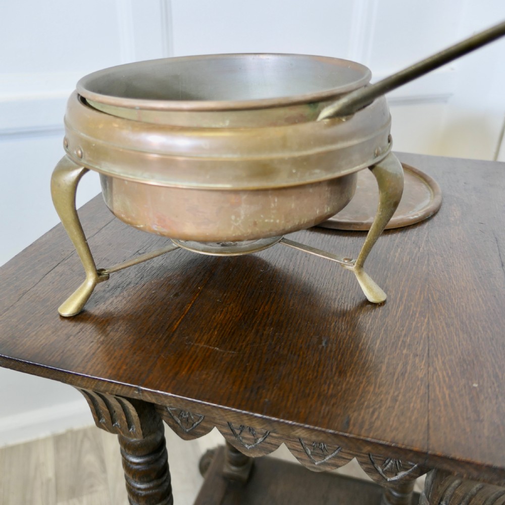 A vintage circular copper bain marie featuring a lid, long handle, and a three-legged brass tripod stand with an integrated burner tray. View II