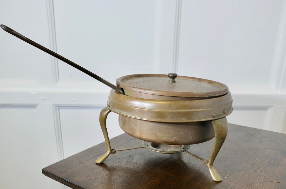 A vintage circular copper bain marie featuring a lid, long handle, and a three-legged brass tripod stand with an integrated burner tray. View V