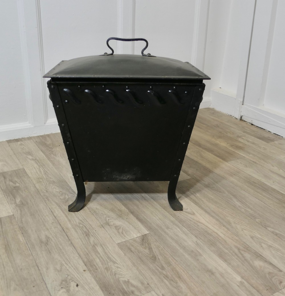 A black riveted metal Arts and Crafts coal box featuring a domed lid with a central handle, decorative wave detailing, and splayed metal legs.